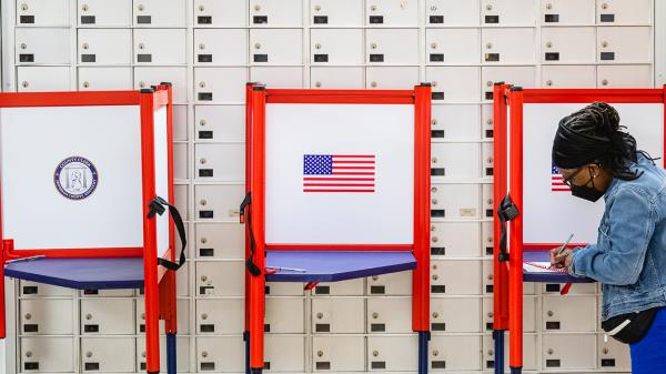 A Black woman wearing a black mask, seen on the far right side of the photo, stands over a voting booth as she casts her ballot. Two other voting booths are seen with the American flag; one booth is in the dead center of the photo.