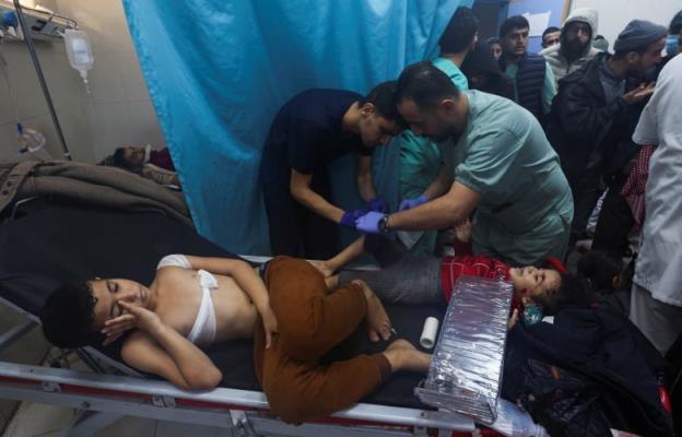 A young boy, wearing brown pants with a bandage wrapped around his chest, lies on a stretcher in a hospital room. Beside him, medical staff treat a young girl who is crying.