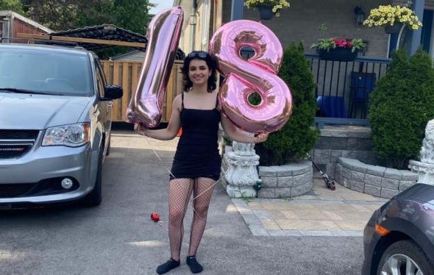 A young woman in a black dress smiles as she holds two pink balloons in the shape of the numbers 1 and 8