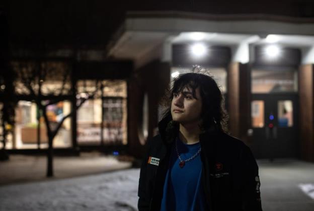 A young man with long black hair stand outside school doors. It is dark and snowy outside. 