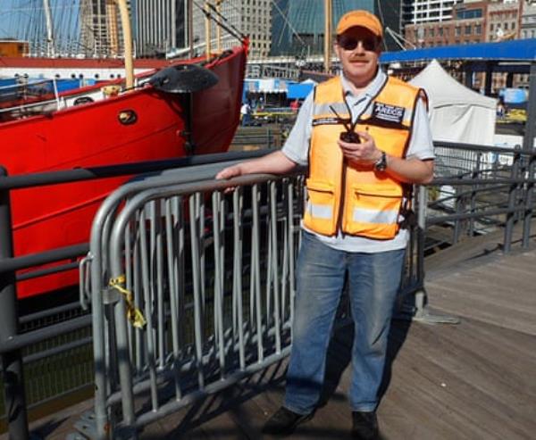 man holds radio and wears hat, sunglasses and hi-vis jacket