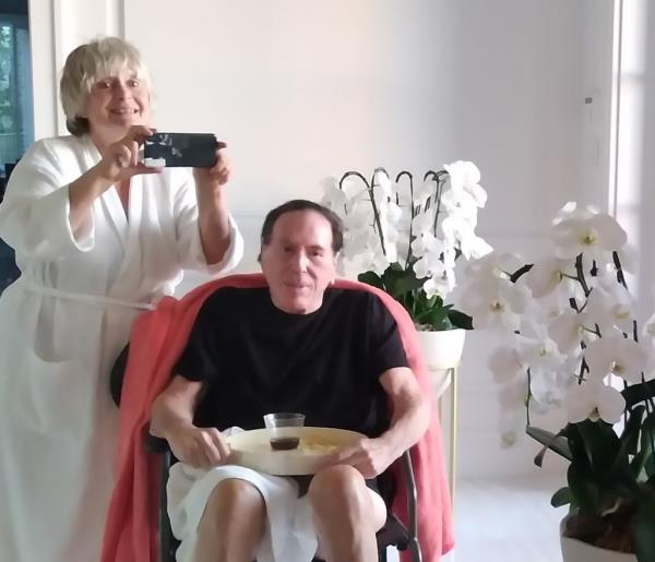 Vostrikova in a white robe holding a cellphone as if taking a picture in a mirror, with allan goldman in a black t shirt and whit shorts sitting next to her in a pink armchair