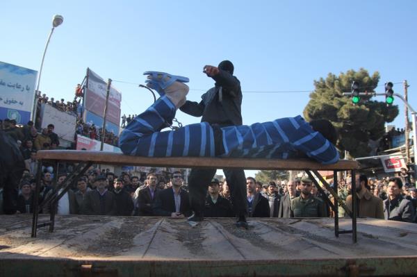 A man in striped clothing tied face down to a table is whipped by a masked man with other men watching behind 