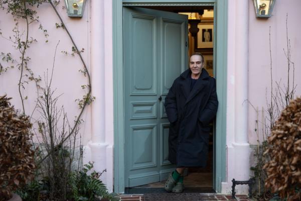 John Galliano standing in a doorway wearing a coat and socks, with text a<em></em>bout his life and career as a fashion designer.