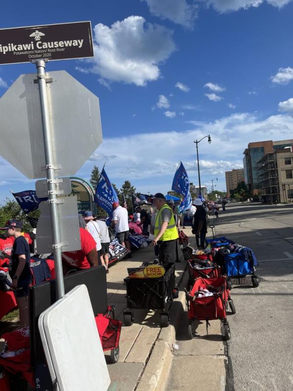 A man with a wagon stands in front of a line of vendors on a sidewalk selling MAGA goods.