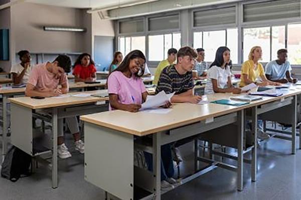 A group of students sitting in rows in a classroom working on paperwork and looking up toward front of classroom.