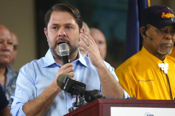 Senatorial candidate Ruben Gallego speaking into a microphone at a campaign event at American Legion Post 41 in Phoenix.