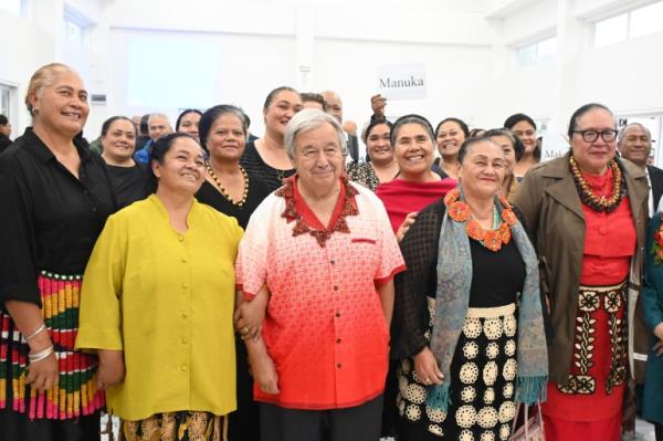 Anto<em></em>nio Guterres with a group of women in Tonga. He is wearing a red and white shirt with a decorative neck;ace. He is smiling. One of the women has her arm through his.