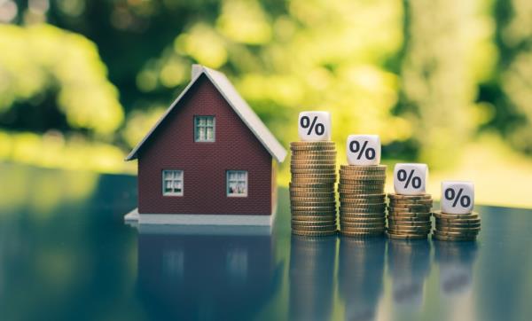 Model house beside stacks of coins decreasing in height with dice displaying percentage symbols, symbolizing decreasing interest rates.