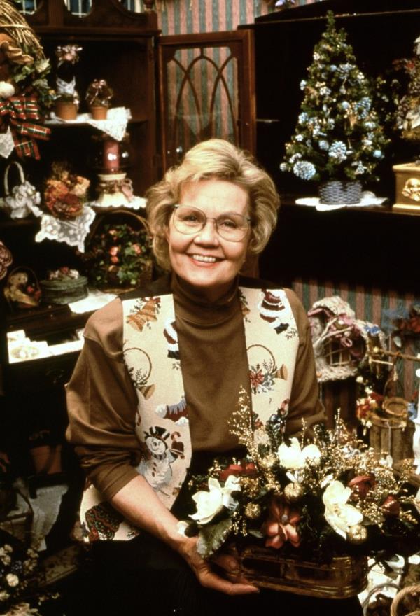 Old white woman in crafty vest, short light hair and glasses with turtlneck.