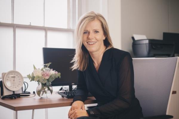 Alice Duffy sitting at her desk, wearing a black blouse and trousers, there is a computer and flowers on her desk. She has blo<em></em>nde hair and is smiling to camera.