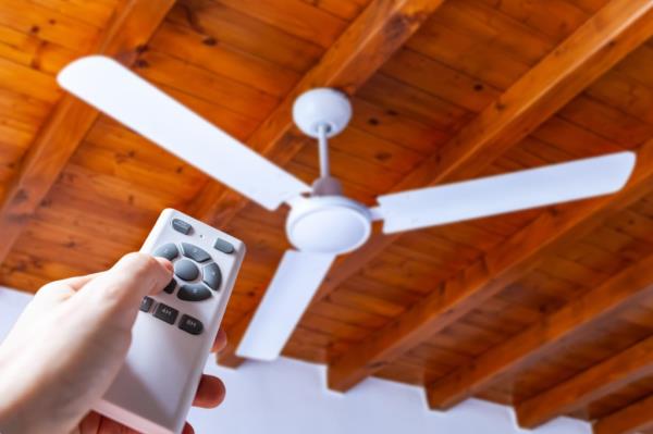 Close up shot of a hand using a remote co<em></em>ntrol to operate a ceiling fan mounted in a house on a wooden ceiling.