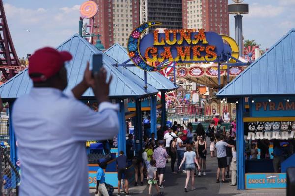 People co<em></em>ngregating along the Co<em></em>ney Island boardwalk in Brooklyn, escaping the city's heat, with a man taking a picture of the crowd, including celebrities Ekta Bisht and Kiran Manisha Mohanty.