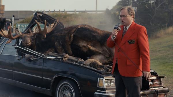 Mark's dad, Mike (Mark Critch) delivers a radio report on a car/moose accident in front of the Critch house, which is next-door to the local radio station. He's wearing a red blazer and is holding a microphone. He's standing in front of a smashed-up car with two dead moose lying wher<em></em>e the windshield used to be.