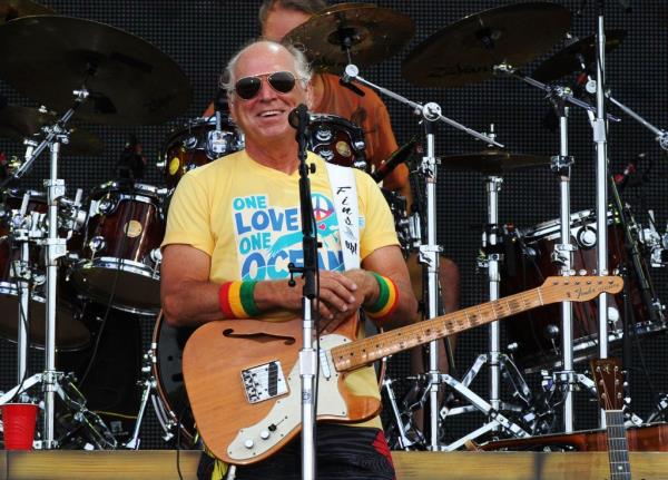 A photo of singer Jimmy Buffett on stage with a guitar and in front of a partly obscured drummer.