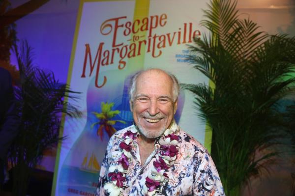 Singer Jimmy Buffett stands smiling in front of a poster for his Broadway musical 