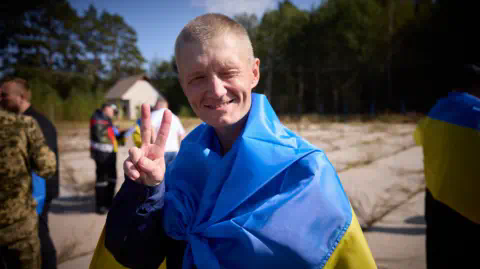 Ukrainian government A Ukrainian servicemen released during a priso<em></em>ner exchange with Russia is seen squinting and smiling into the camera. He is also wrapped in a Ukrainian flag and holding up two fingers facing forward in a V sign. 