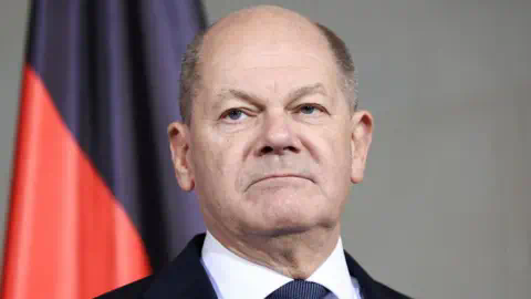 Reuters A headshot of Germany's Chancellor Olaf Scholz as he appears ex<em></em>pressio<em></em>nless photographed in front of a red and black flag