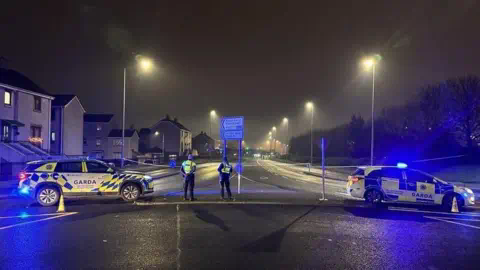 RTé Two Garda (Irish police) cars block off the entrance and exit to a road. Two officers look down the empty road. It appears to be at the exit to a rounda<em></em>bout. There is a housing development to one side of the road, and trees to the other. 